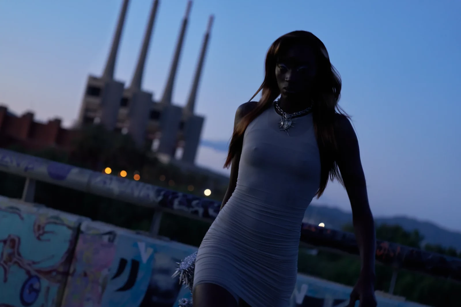 Full-length portrait of a dark-haired model in a fitted pale blue dress standing by the water at dusk.