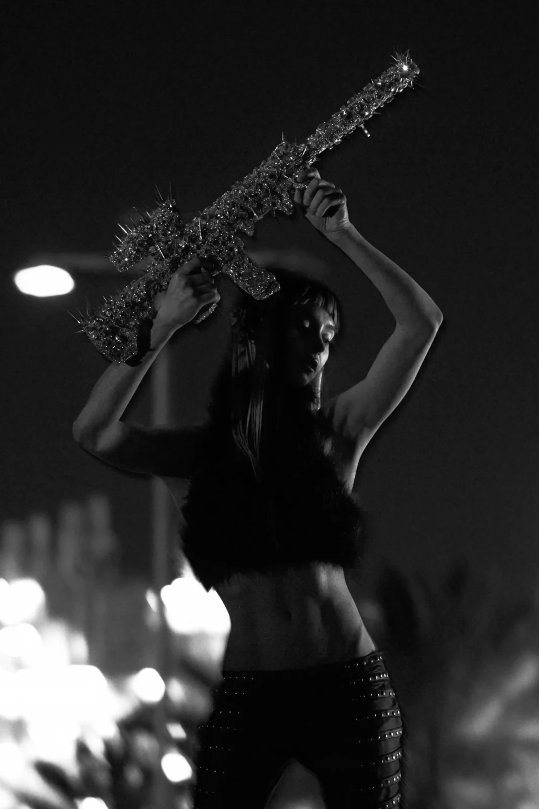 Black-and-white photograph of a model holding a large textured prop above her head at night.