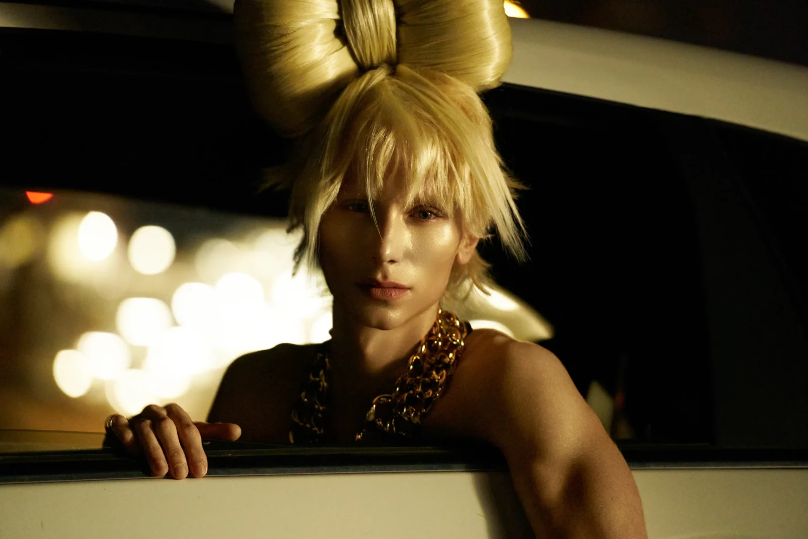 Blonde fashion model with sculptural hair buns leaning out of a car window under warm streetlights.