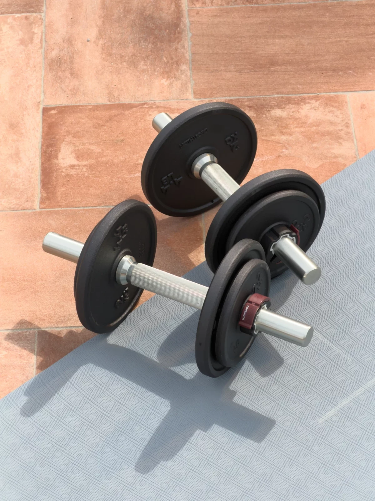 Pair of adjustable black dumbbells and weight plates arranged on pavement beside a terracotta wall.