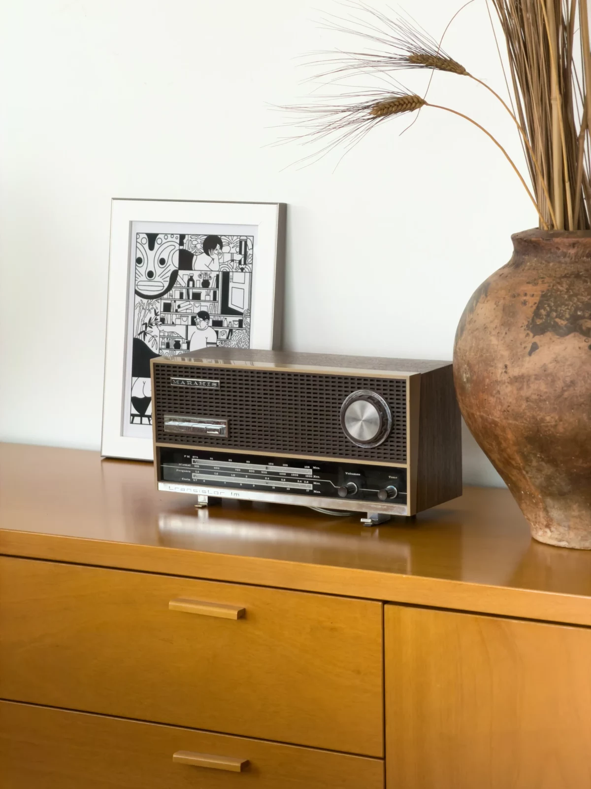 Vintage black radio displayed on a wooden sideboard beside a framed print and a tall ceramic vase.