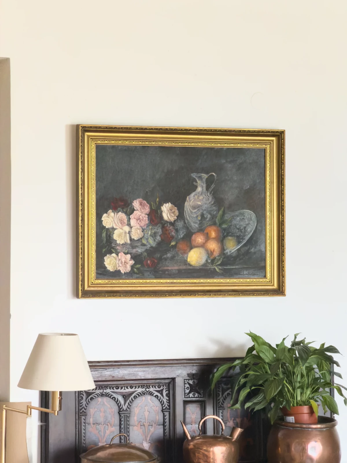 Framed still-life painting of fruit above a wooden cabinet, cream lamp and houseplant.