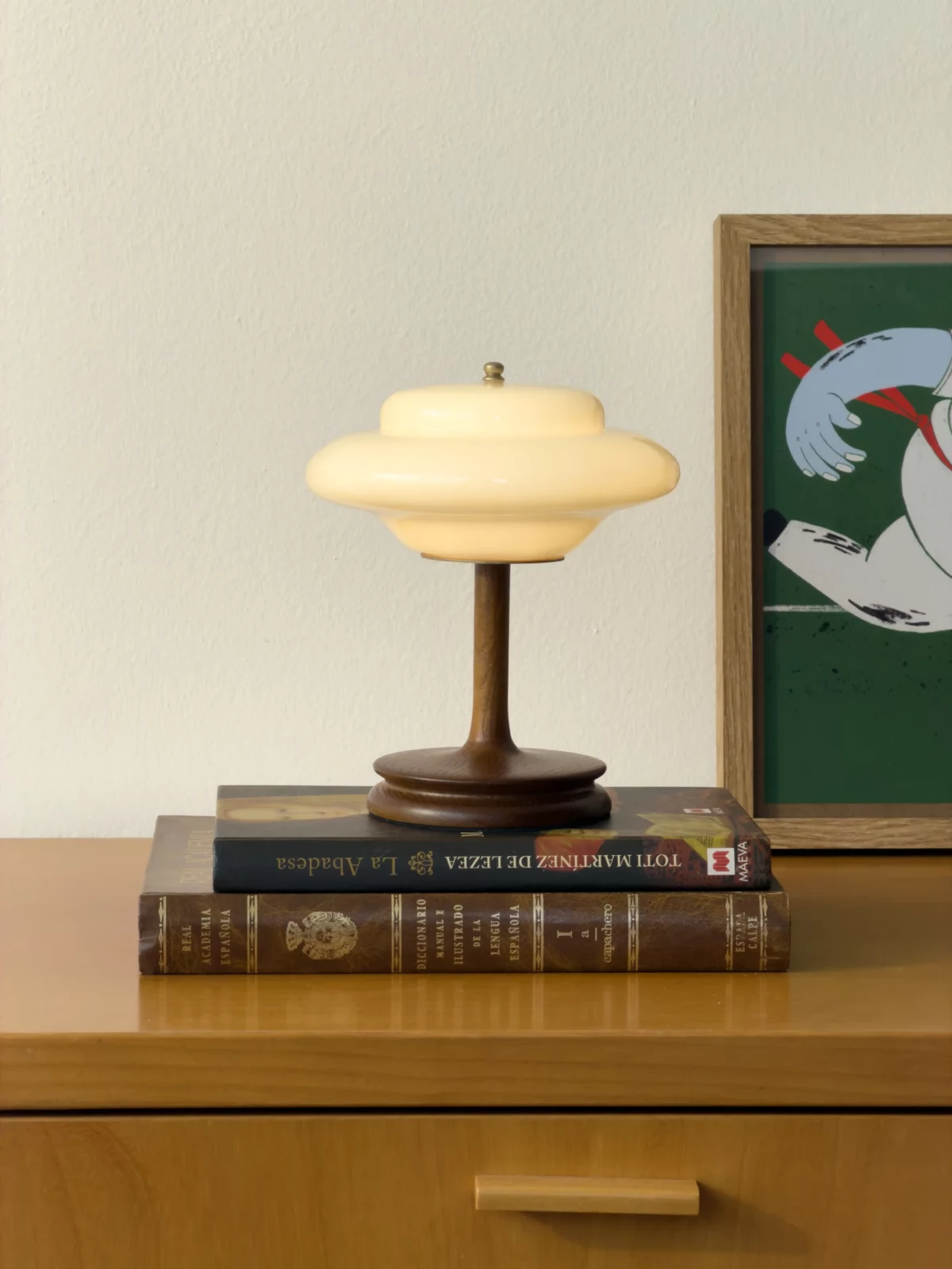Small cream-colored table lamp placed on stacked books beside framed artwork.