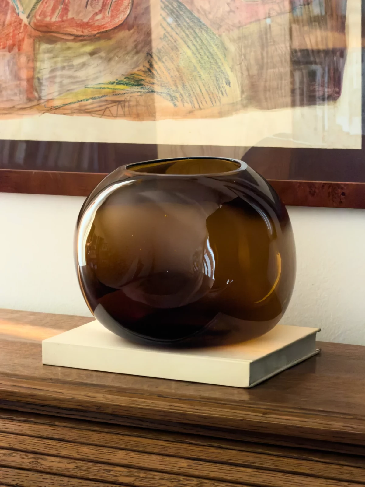 Rounded dark glass decorative vase resting on stacked books in front of a framed artwork.