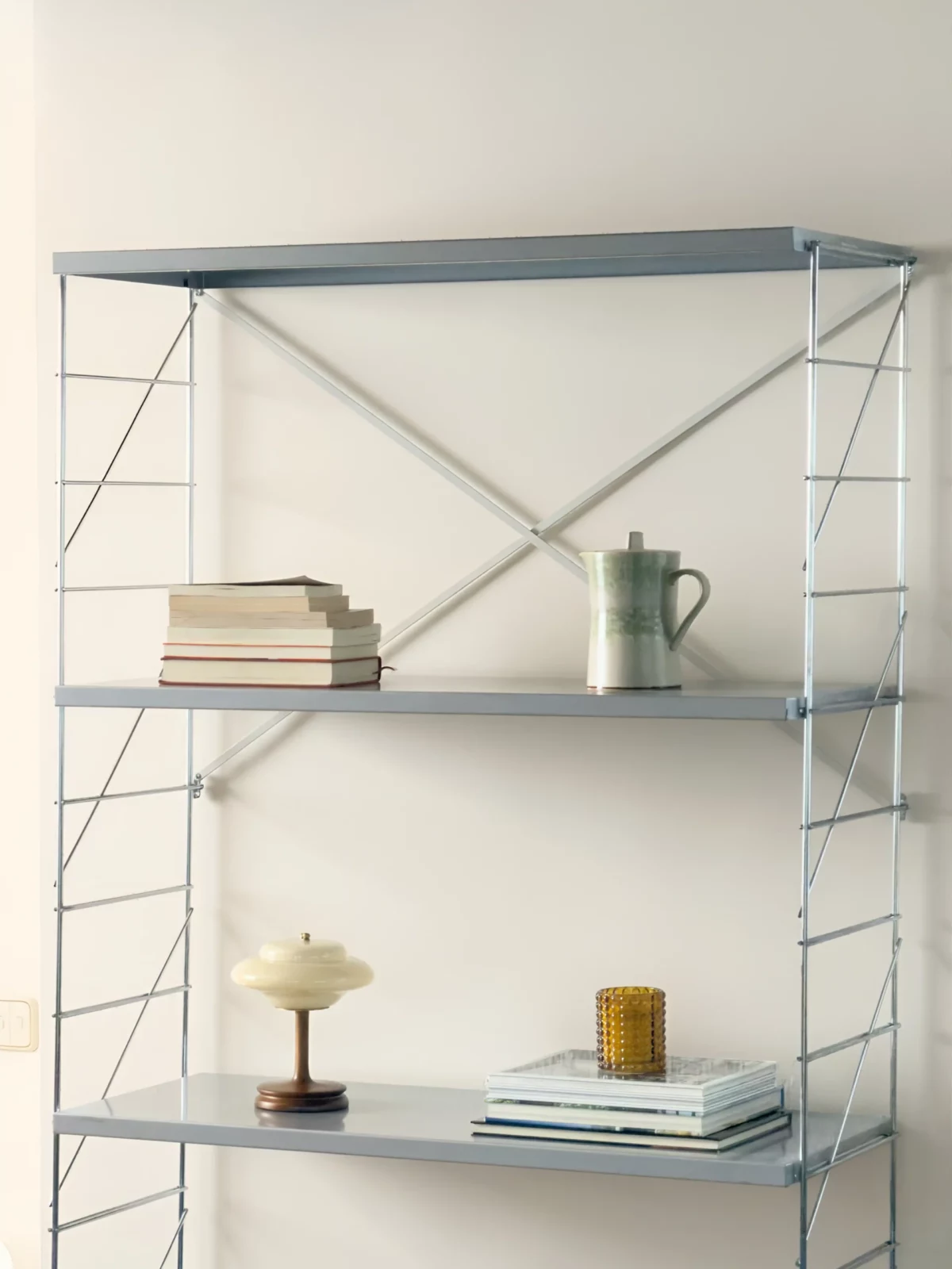 Tall open metal shelving unit displaying books, small plants and decorative objects against a white wall.