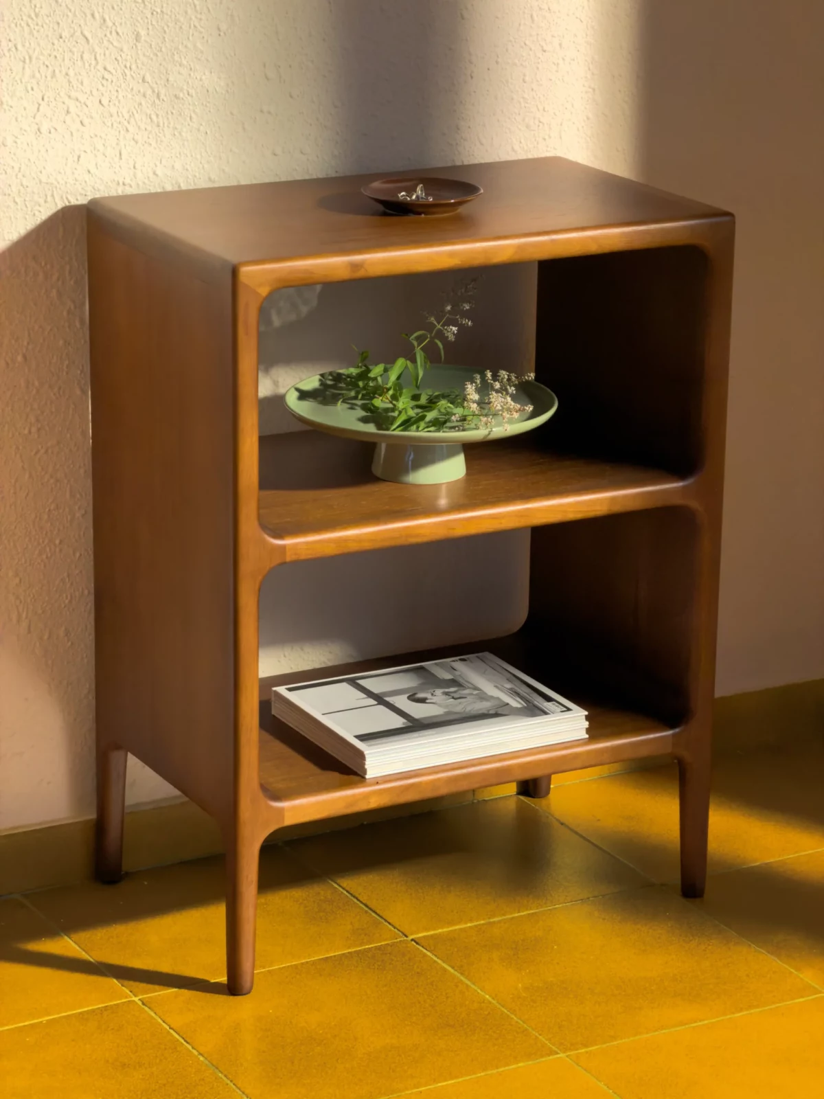 Small wooden bedside cabinet with open shelves, books and a bowl of green plants.
