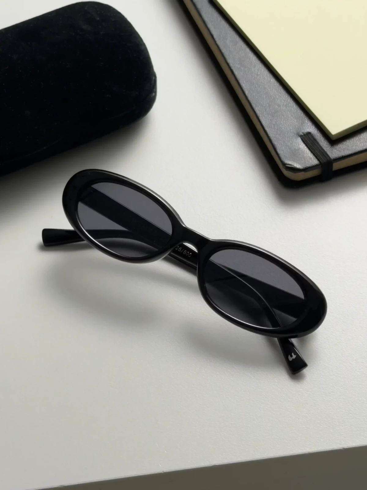 Black oval sunglasses resting beside a dark case and a tablet on a light desktop.