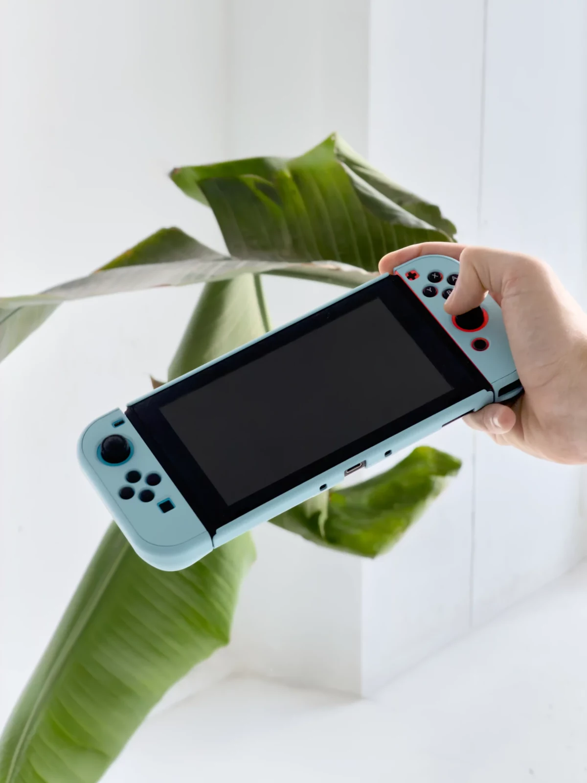 Hand holding a pale blue portable gaming console in front of broad green plant leaves.