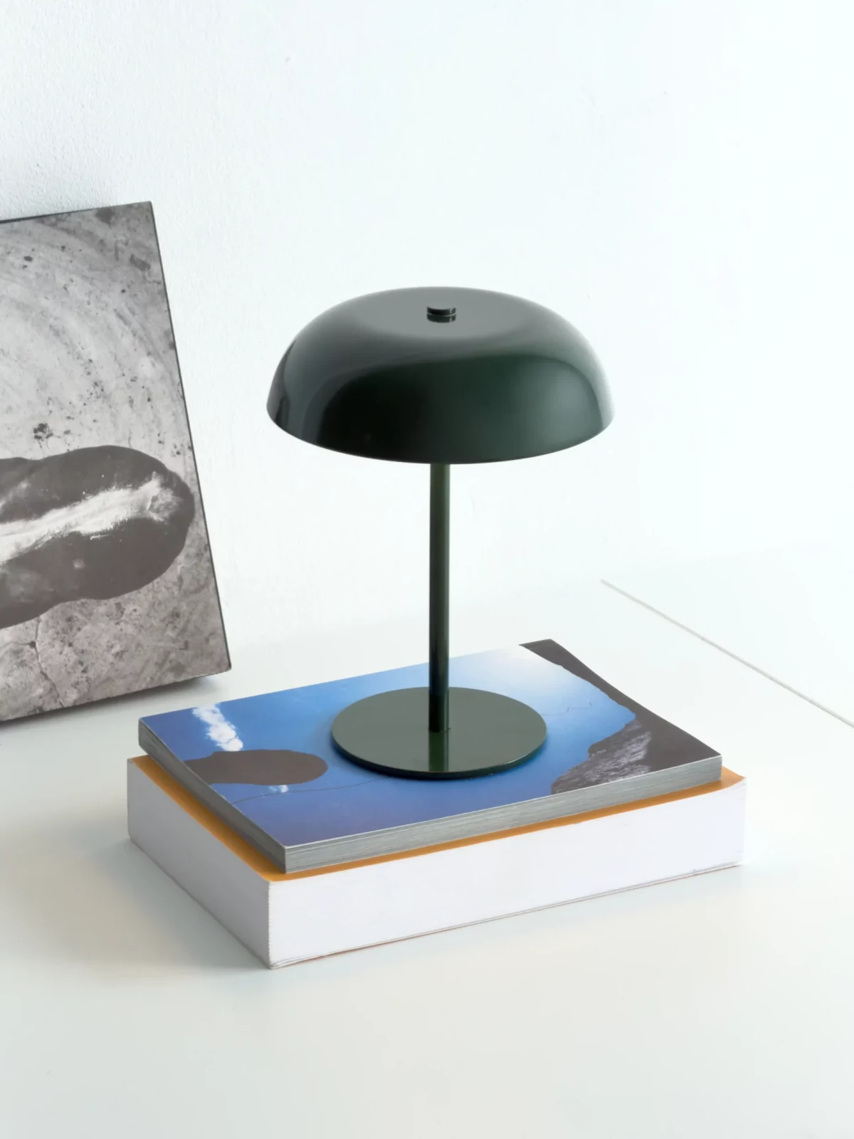 Dark green mushroom-shaped table lamp displayed on top of a blue book against a light wall.