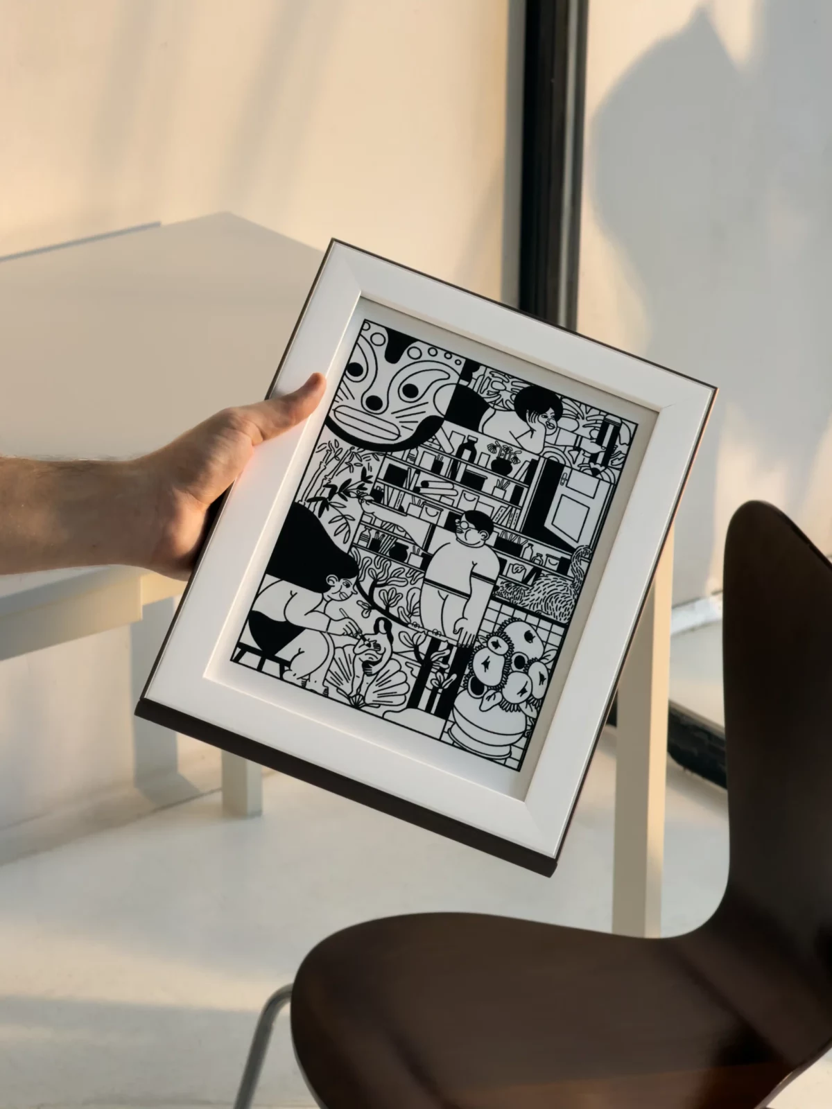 Person holding a framed black-and-white illustration in a bright minimalist interior.