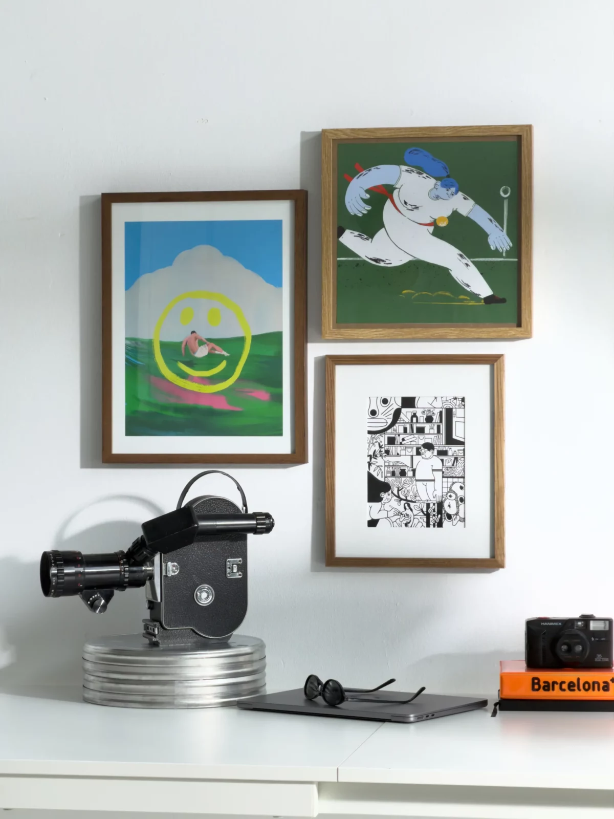 Group of colorful framed prints hung above a small coffee machine and books on a white wall.