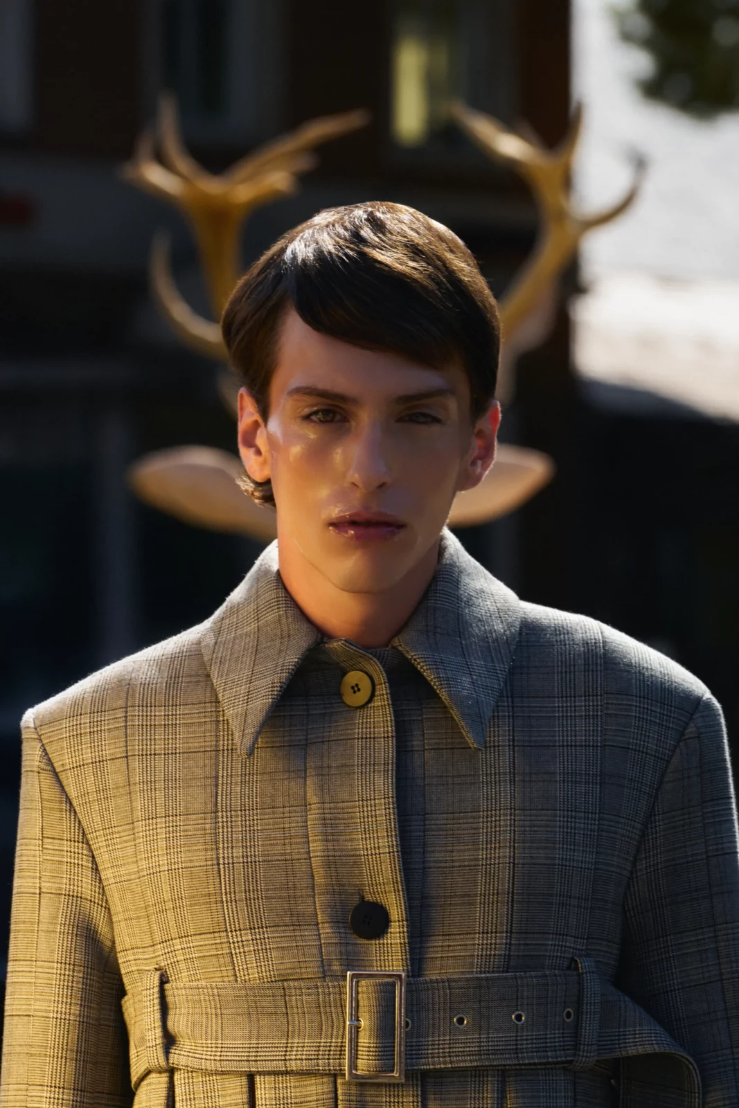 Male fashion model in a gray plaid shirt posing with a deer sculpture behind him.