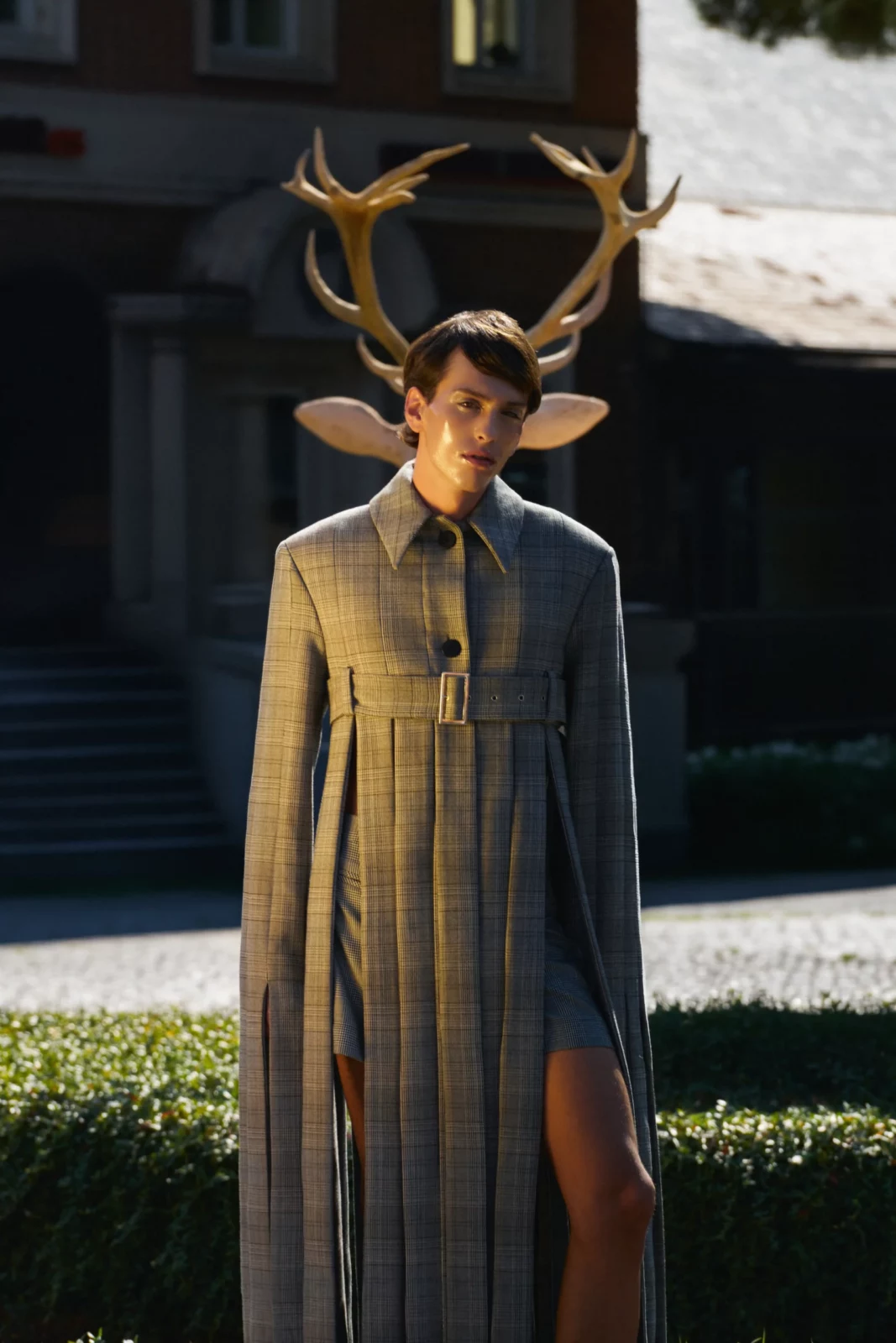 Full-length fashion portrait of a model in gray tailored clothing standing before a large deer sculpture.