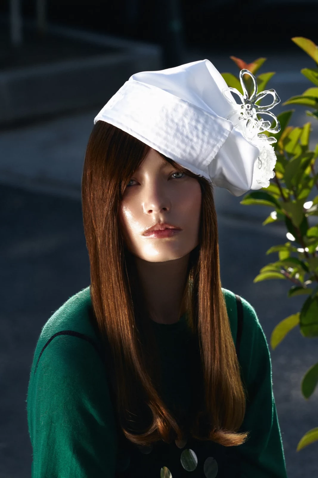 Woman with long hair and a white sculptural hat wearing a green outfit outdoors in warm sunlight.