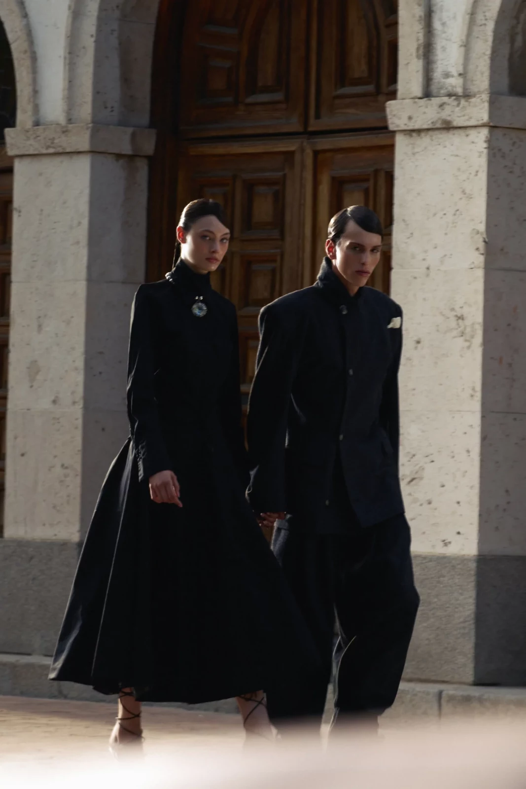 Two models in long black tailored outfits walking in front of an arched stone doorway.