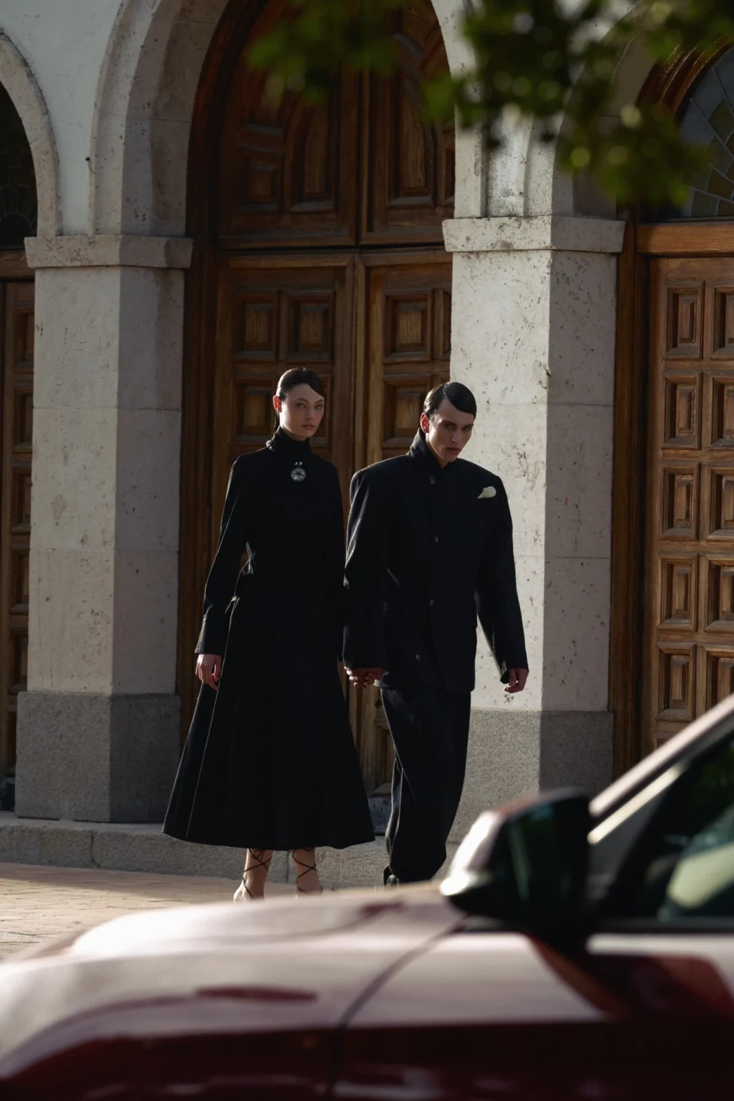 Pair of models in black clothing walking through a colonnade, partly obscured by a passing vehicle.