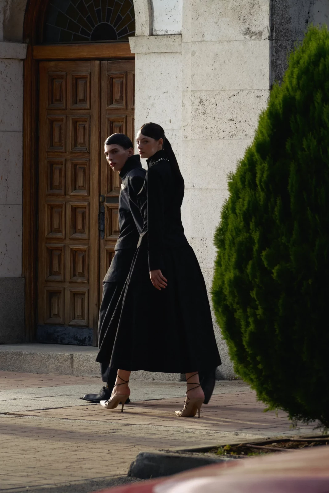 Side view of two models wearing black coats walking alongside a historic stone building.