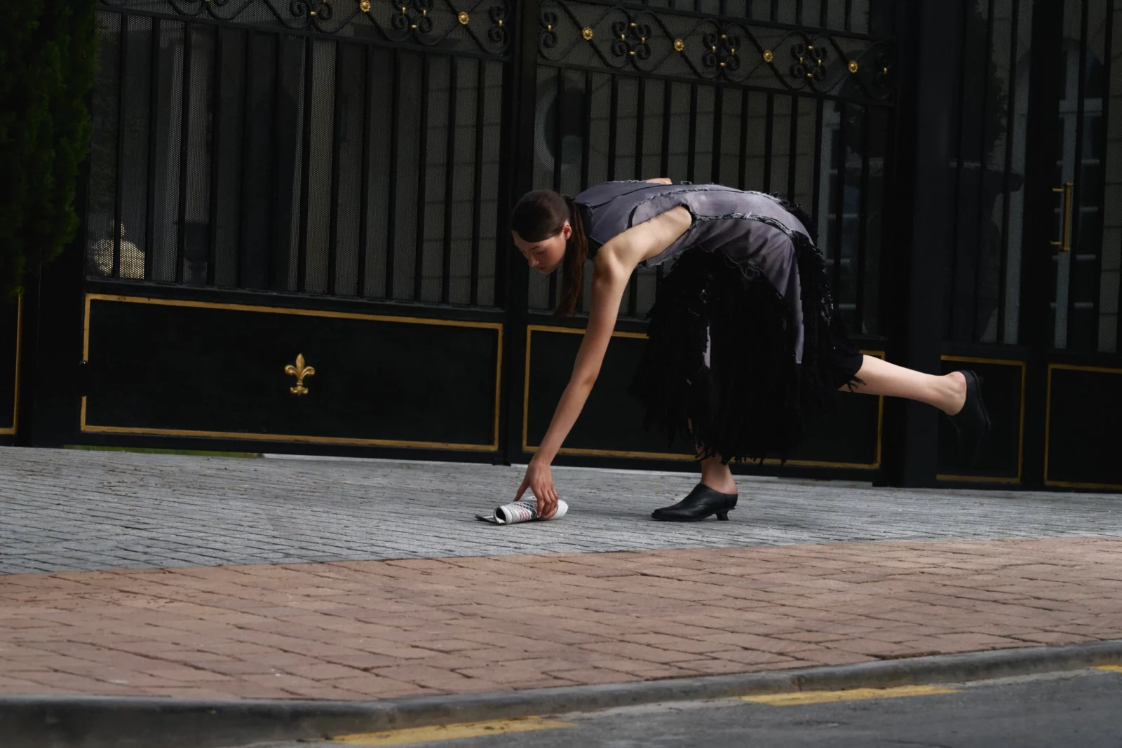 Model wearing a gray and black outfit crouching mid-step on a cobbled street by ornate gates.