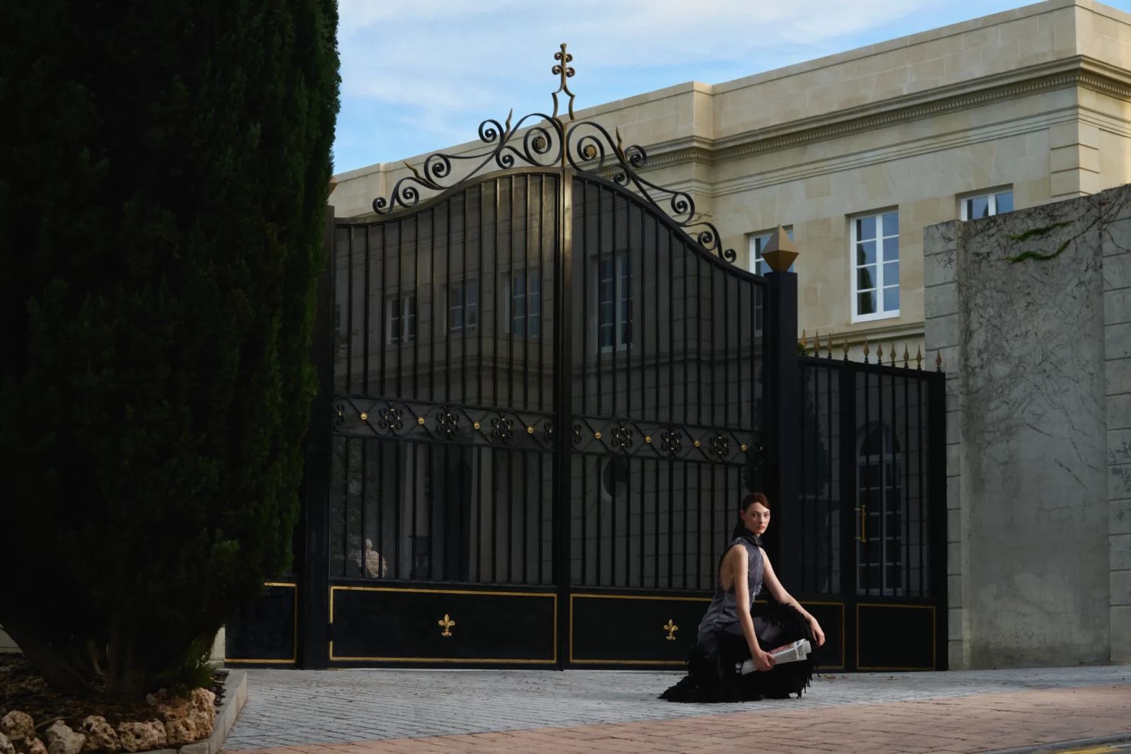 Fashion model in a dark sleeveless dress sitting alone on pavement beside tall black iron gates.