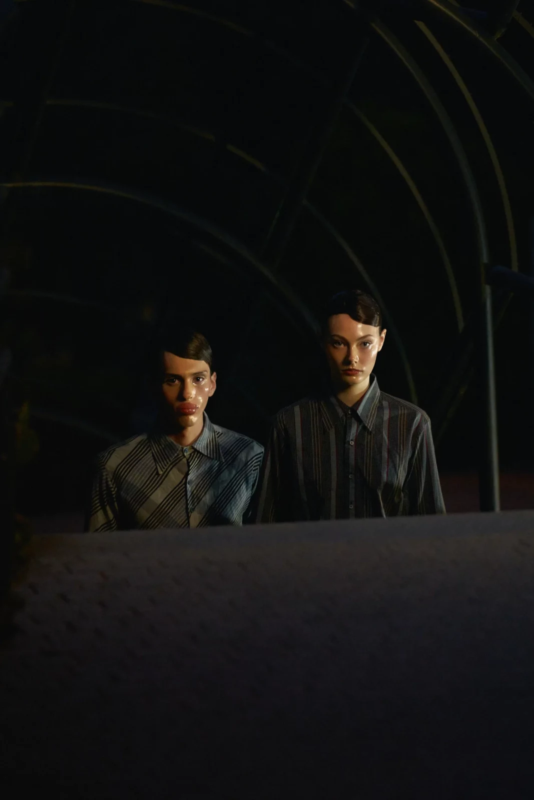 Two fashion models in dark tailored outfits framed in shadow behind a curved stone opening.
