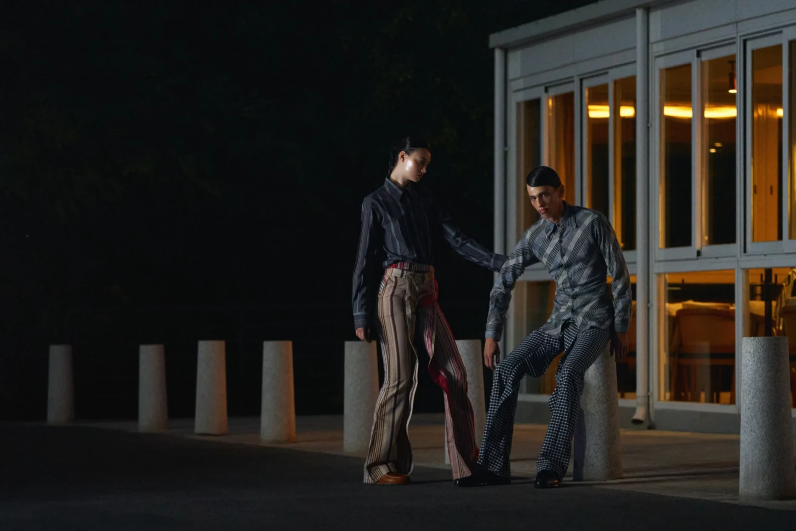 Two models in contemporary tailored clothing standing outside a modern glass doorway at dusk.