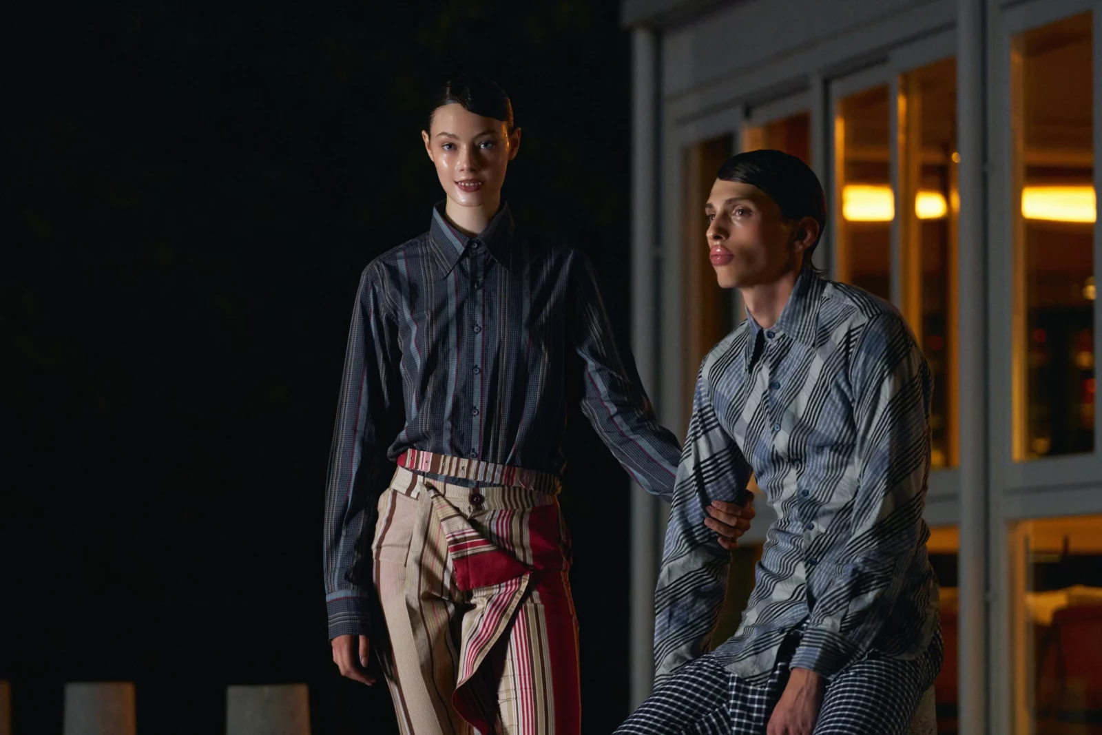 Two models wearing striped and dark shirts posing together against a glass-fronted building at night.