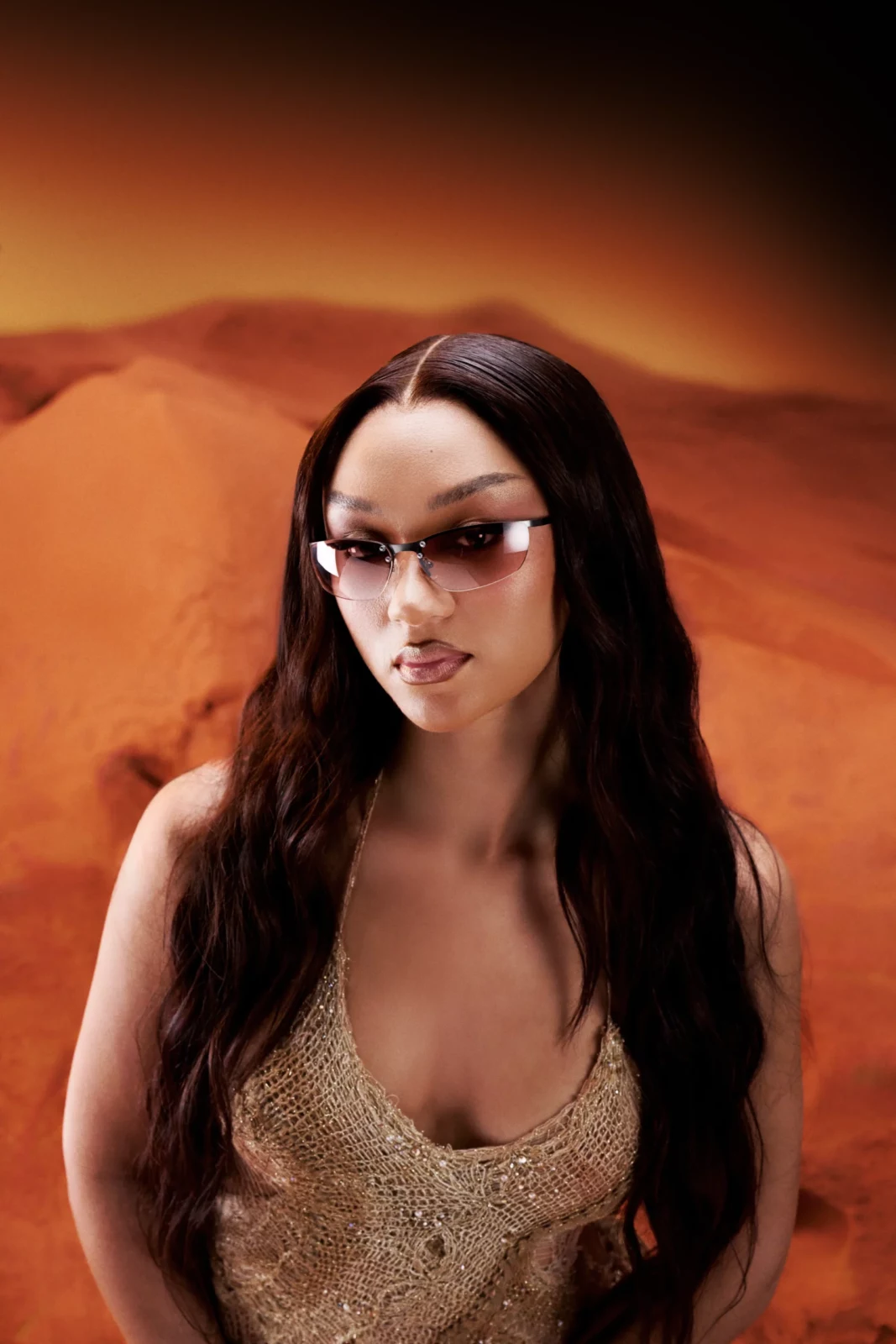 Close fashion portrait of a FLO singer in a pale beige outfit and narrow sunglasses against an orange landscape.