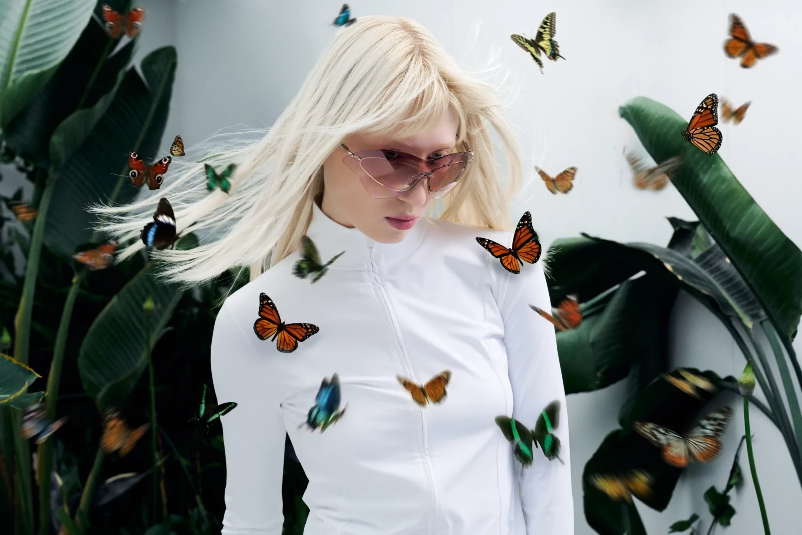 Blonde model in a white high-neck top and pink sunglasses surrounded by tropical leaves and butterflies.