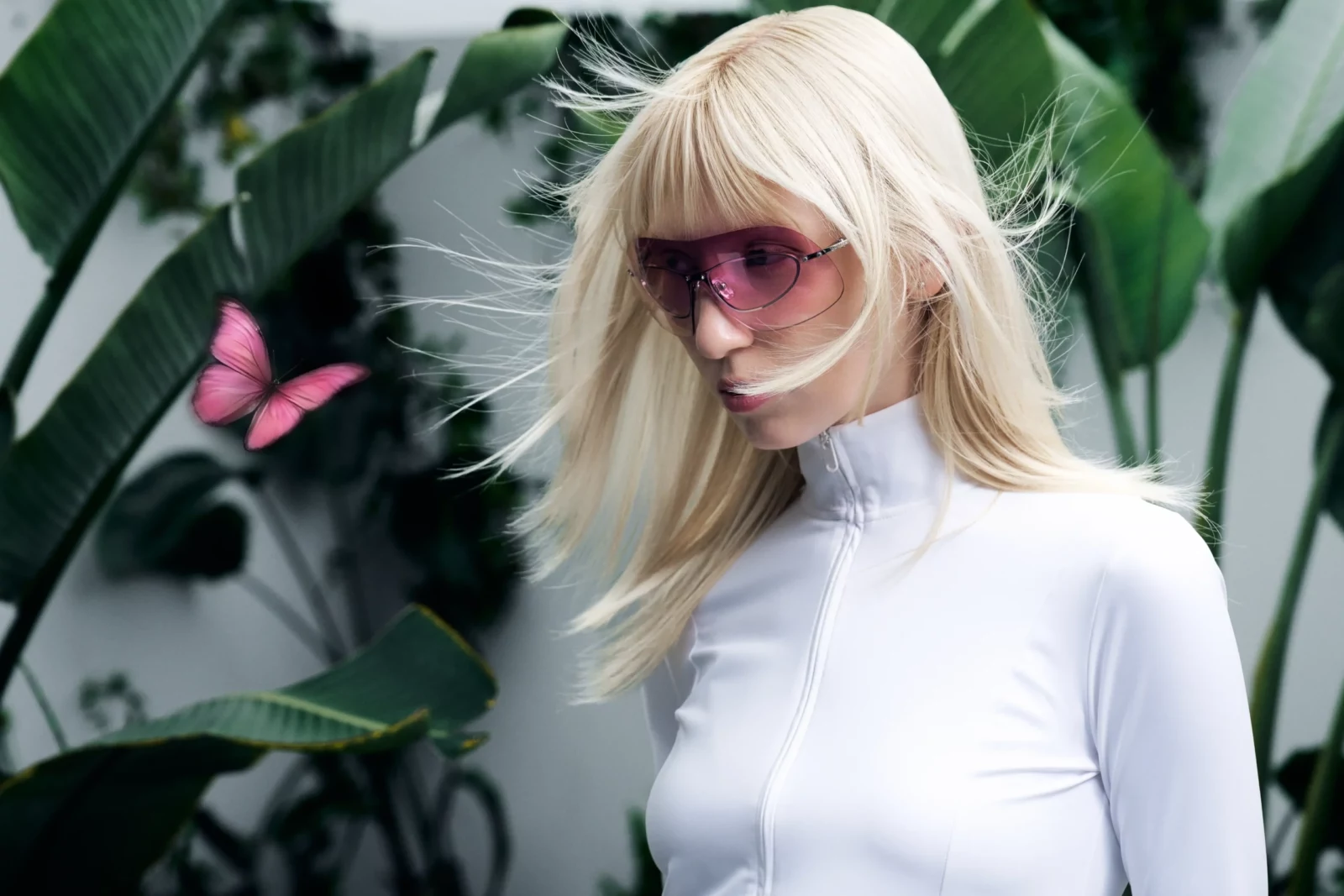 Close-up of a blonde model in pink sunglasses and a white top among lush green foliage.