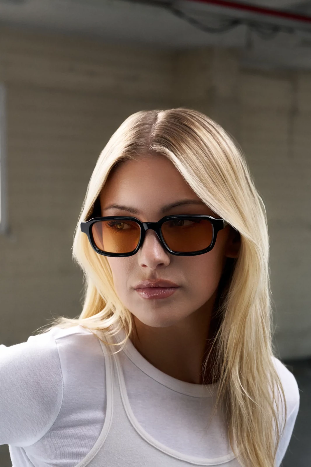 Blonde model in rectangular brown sunglasses and a pale outfit against a concrete background.
