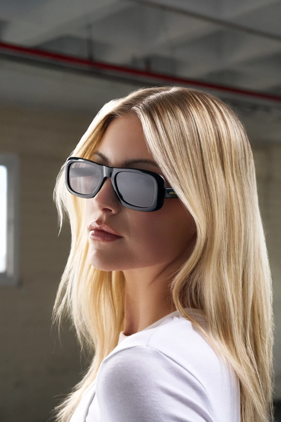 Blonde woman wearing silver framed sunglasses photographed against an industrial interior.