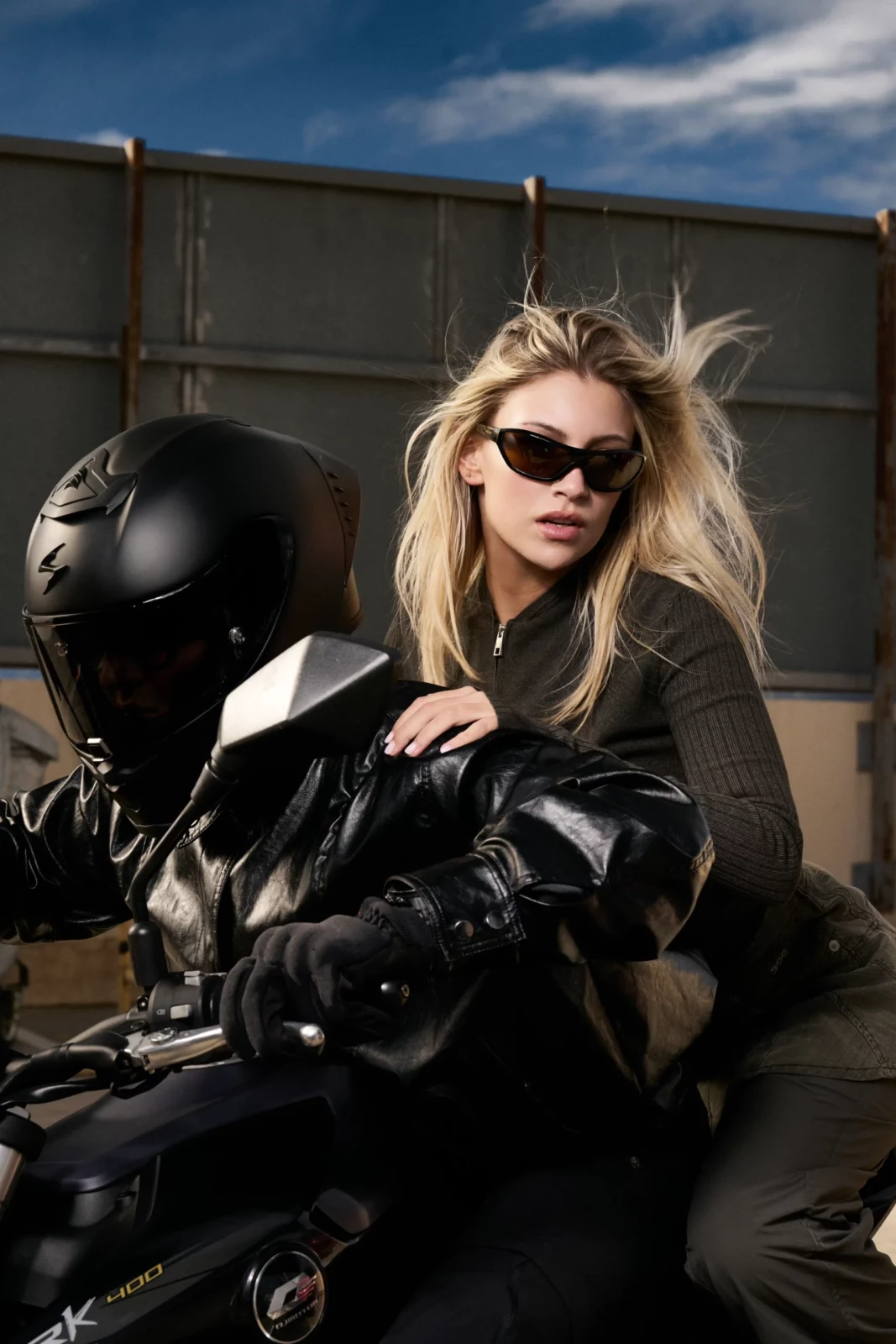 Blonde model in black sunglasses and a dark jacket surrounded by photographers outdoors.