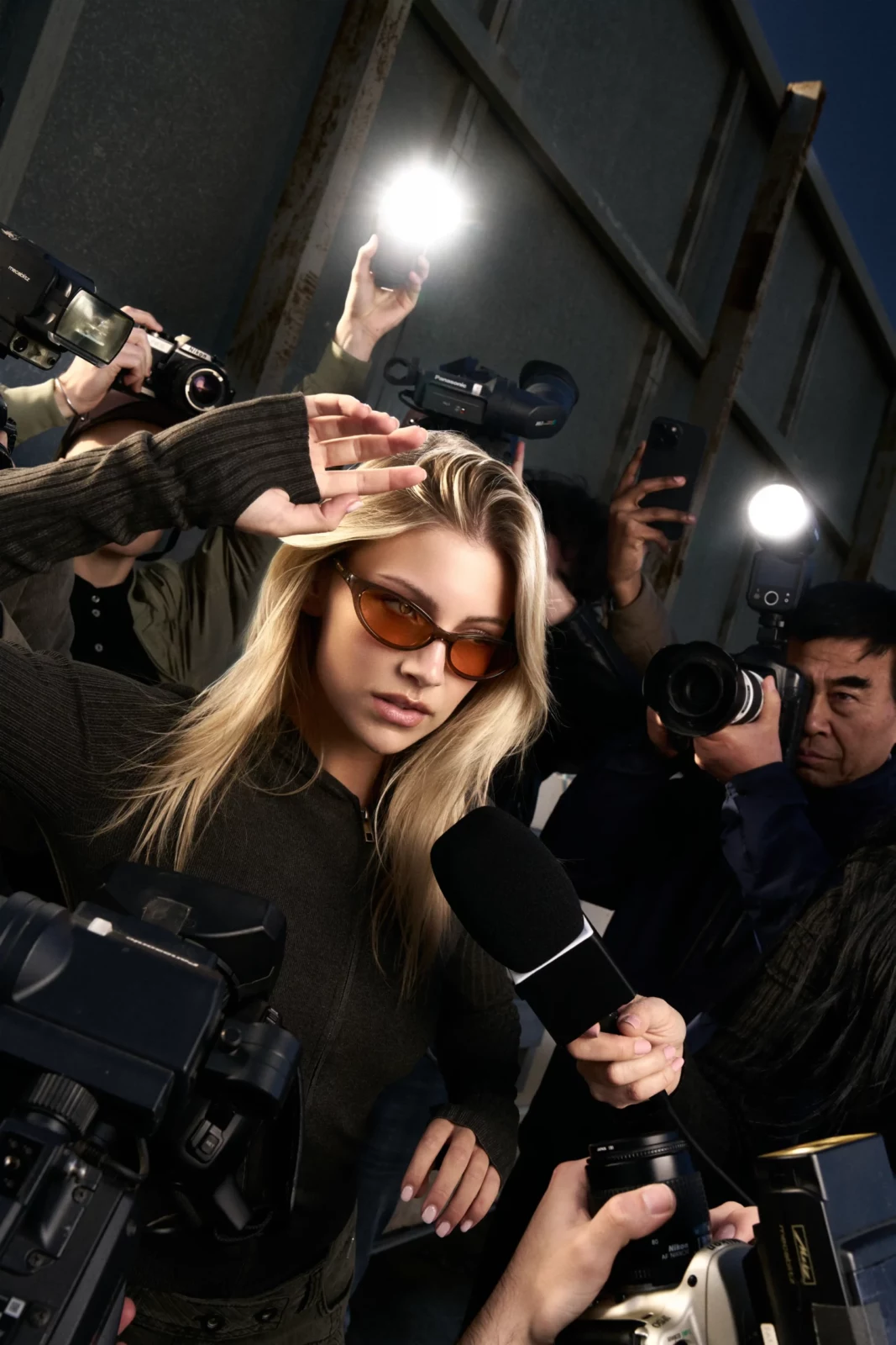 Blonde model in sunglasses surrounded by photographers and bright camera flashes outdoors.