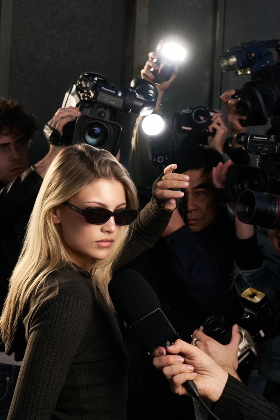 Close-up of a blonde model in black sunglasses with people and cameras behind her at night.