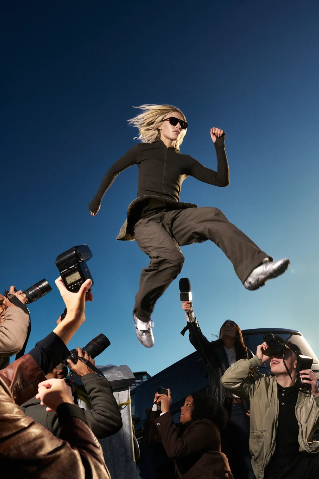 Fashion model in sunglasses jumping in the air above a crowd outdoors under a blue sky.