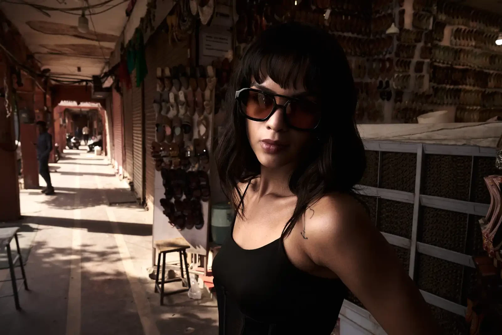 Dark-haired model wearing black sunglasses and a sleeveless top in a sunlit stone passage.