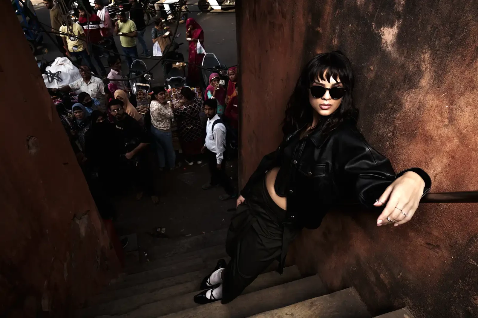 Woman in black sunglasses leaning over a railing above a crowd on a sunlit city street.