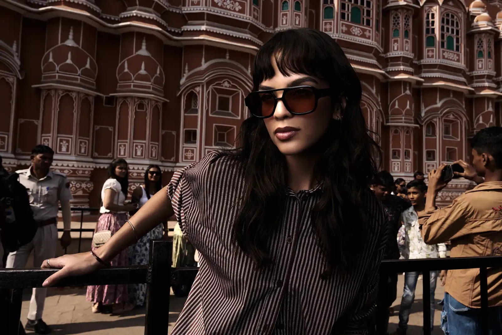 Woman with dark hair and sunglasses standing in front of richly carved stone architecture.