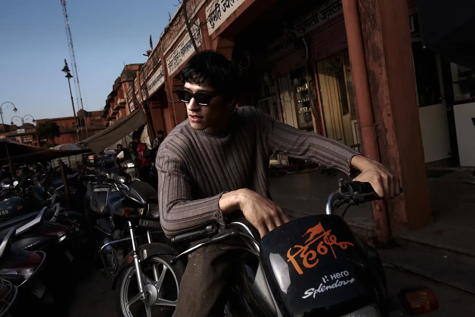 Model in a sleeveless gray outfit and dark sunglasses leaning on a motorcycle on an urban street.