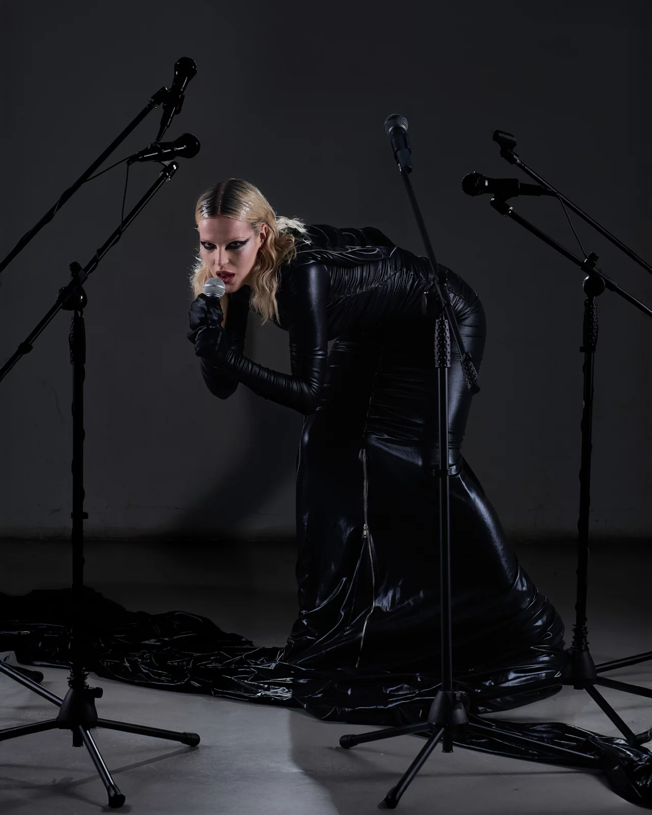 Blonde performer in black leather crouching among microphone stands on a dark stage.