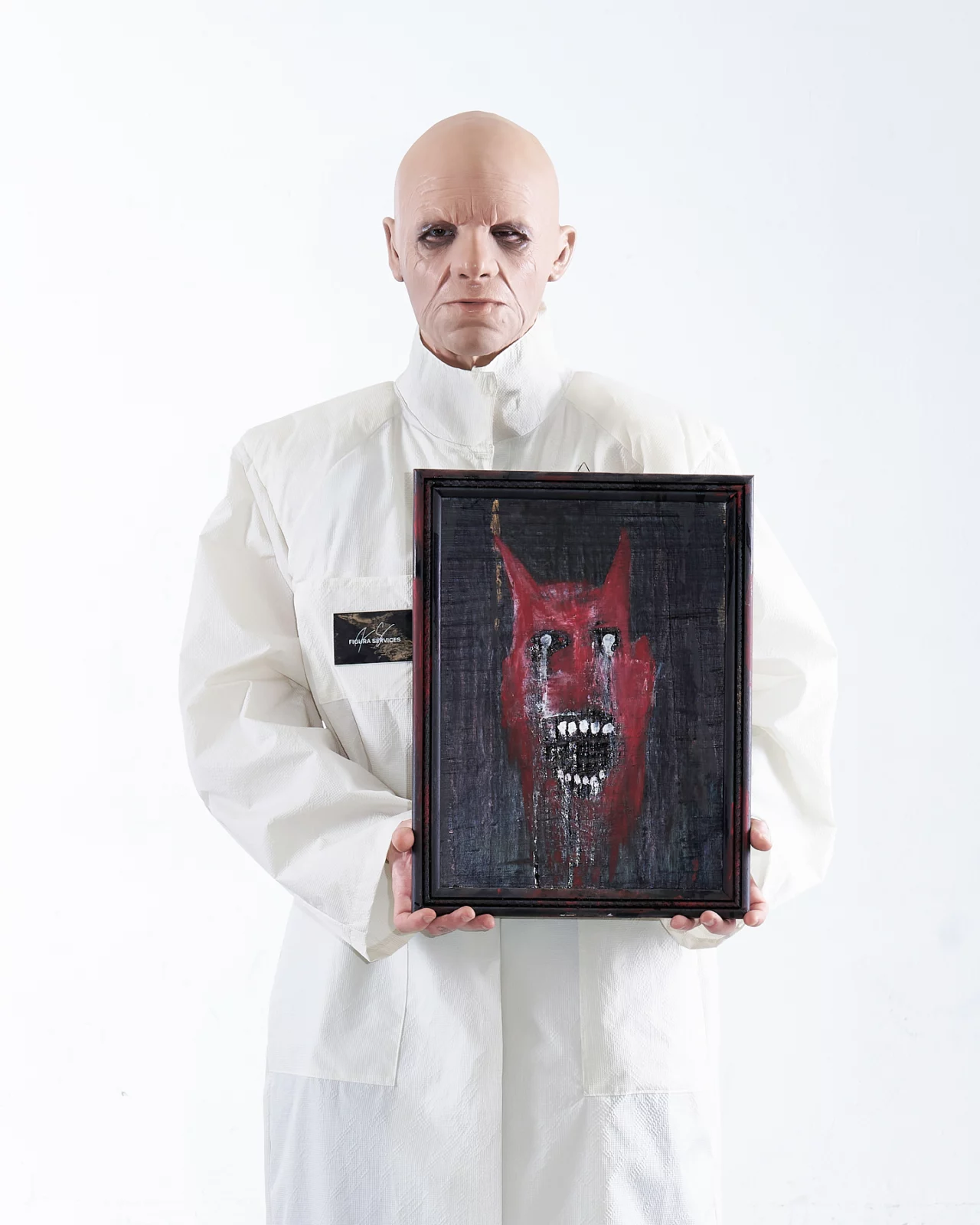 Bald model dressed in white holding a framed dark red artwork against a pale backdrop.