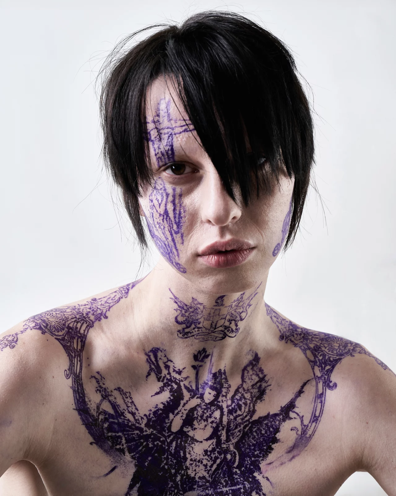 Portrait of a model with black hair, dark lipstick and purple markings across their face and chest.