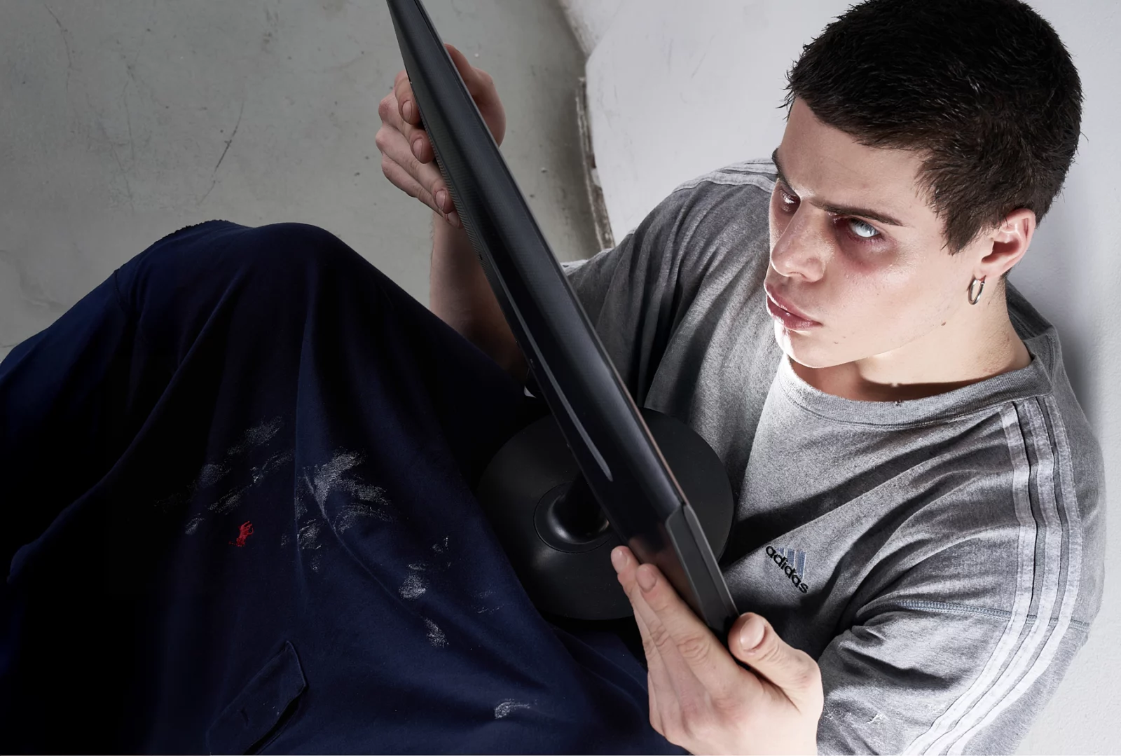 Short-haired model in a gray outfit holding a long black object while seated on the floor.