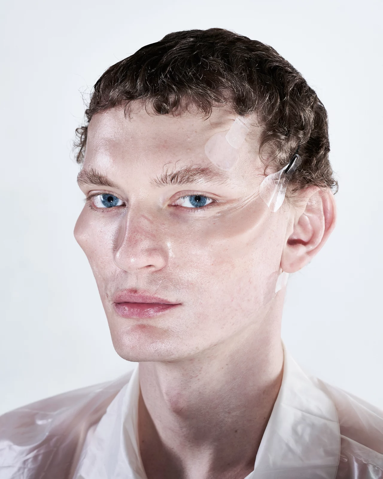 Close-up portrait of a short-haired model with glossy skin and pale eyebrows against white.