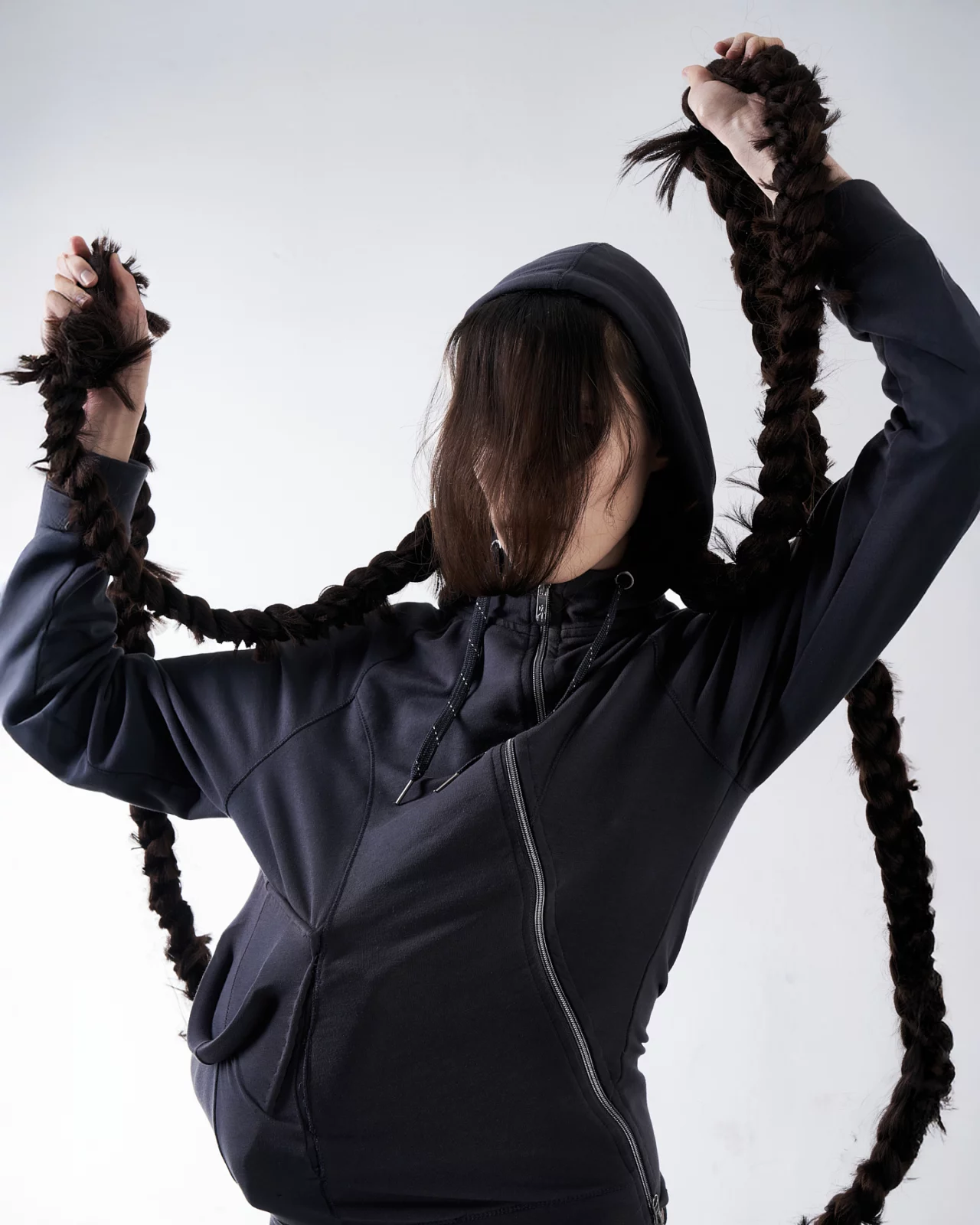 Back view of a model in a black hooded outfit raising both hands and holding long braided hair.